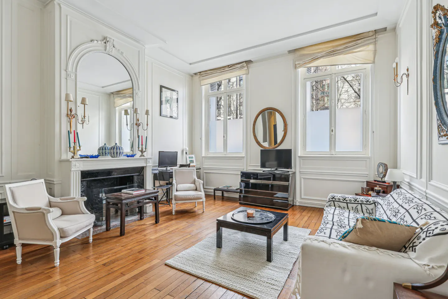 Bright living room with hardwood floors, a fireplace, white armchairs, a patterned sofa, and large windows.