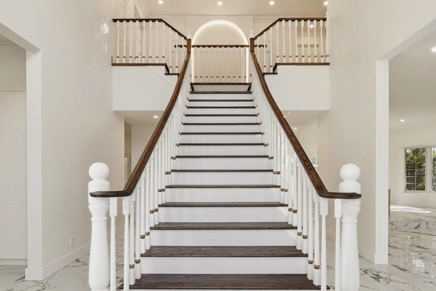 Grand foyer with a staircase. White walls, marble floors, and dark wood stairs with white railings. Natural light fills the space.