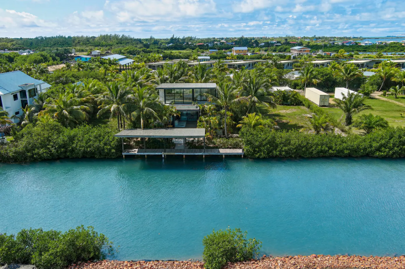 Aerial view of a modern two-story house with a dock on a turquoise canal, surrounded by lush green trees.
