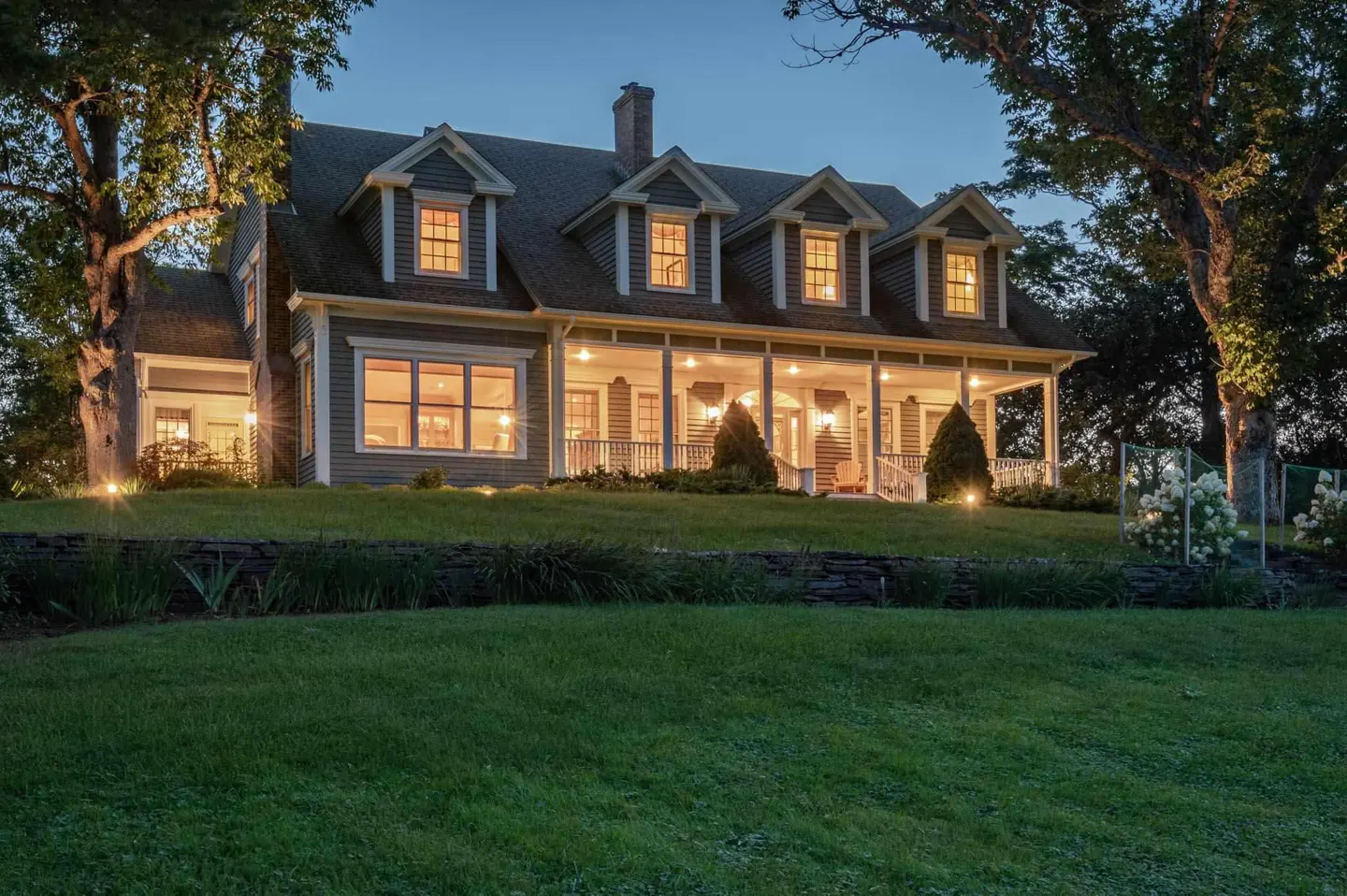 Twilight view of a gray two-story house with a white porch, dormers, and a green lawn.