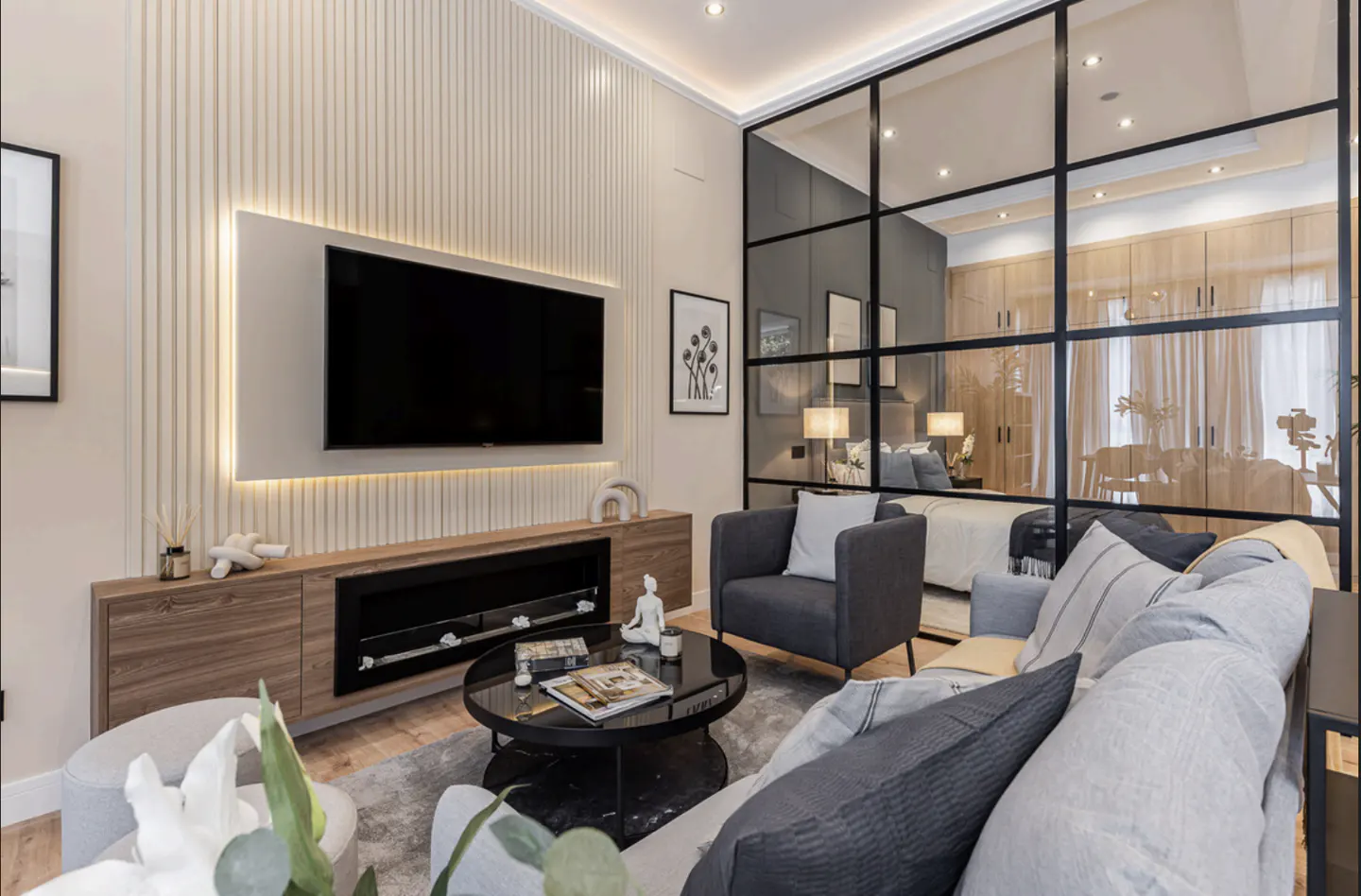 Living room with a black TV above a wooden cabinet, a black coffee table, and a gray sofa, separated from the bedroom by a glass wall.