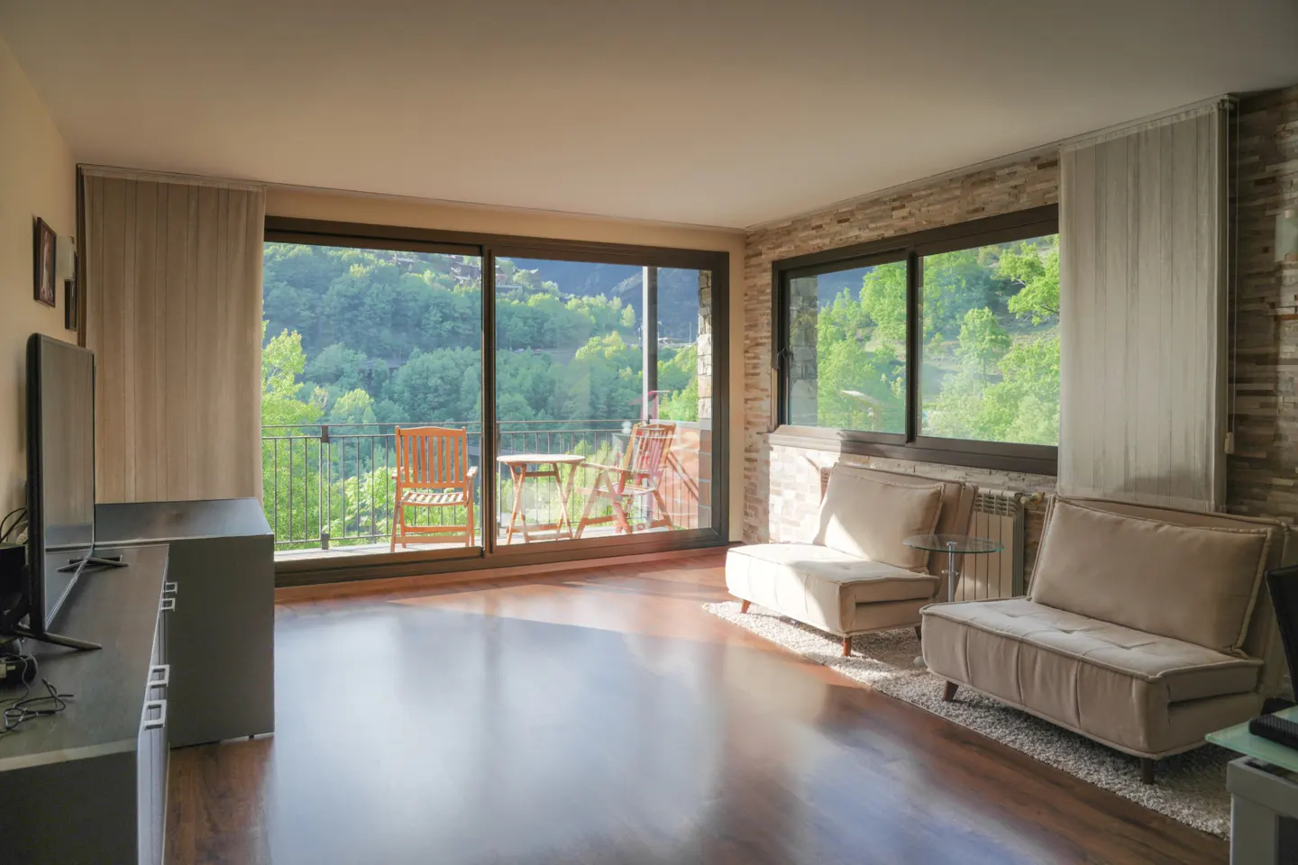 Living room with wood floors, stone accent wall, and sliding glass doors to a balcony with forest views. Two beige sofas sit on a rug.