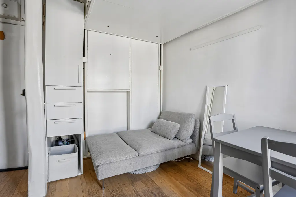A small, all-white studio apartment with a gray chaise lounge, table, and chairs.
