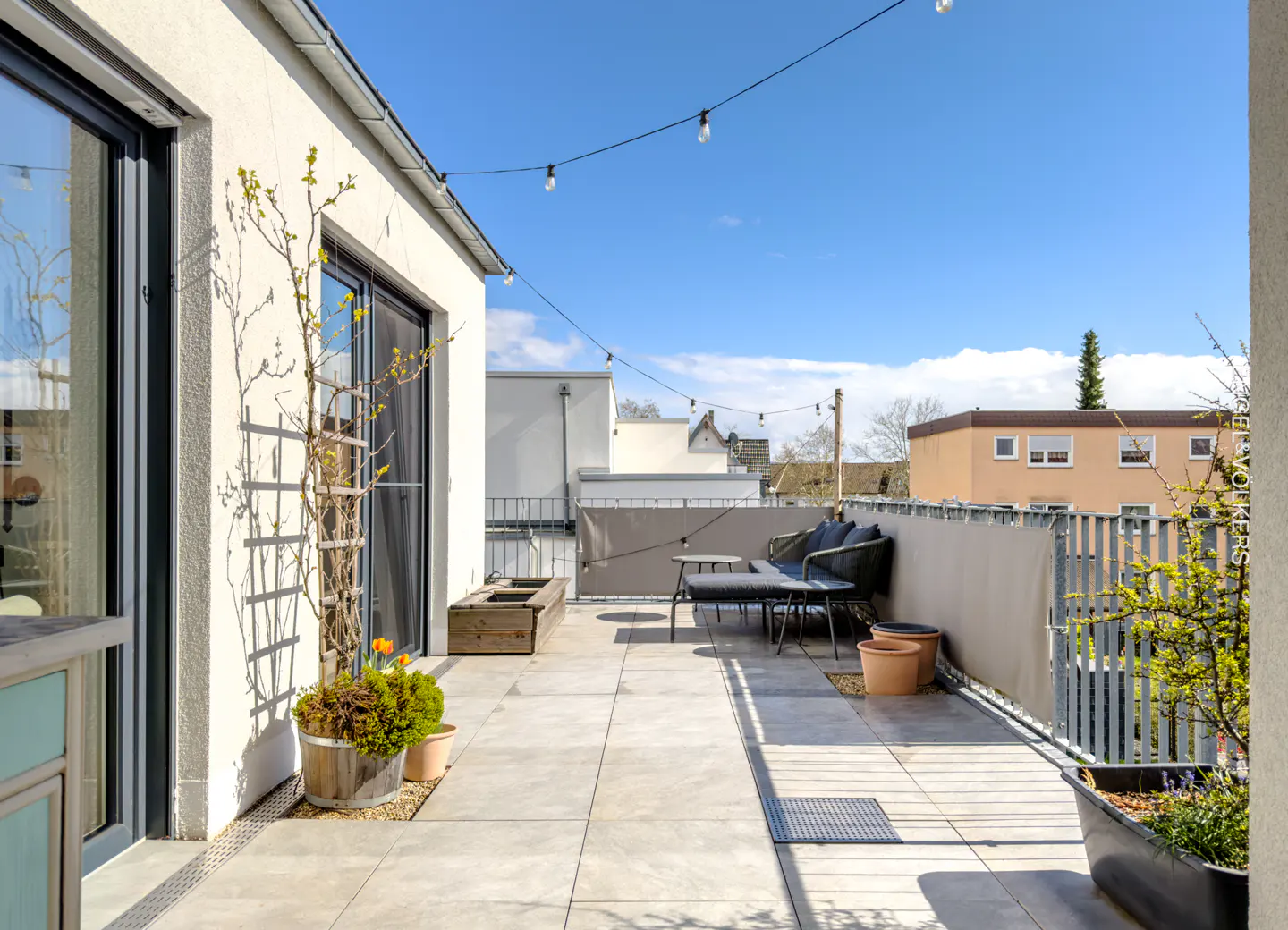 Rooftop patio with gray tile, outdoor seating, and string lights. Potted plants add greenery, with buildings visible in the background.
