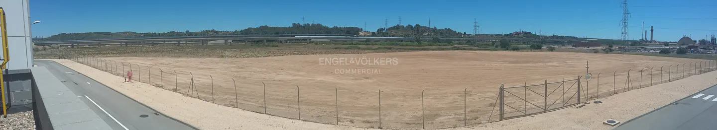 Fenced, vacant dirt lot next to a road, with a highway overpass and industrial structures in the background under blue sky.