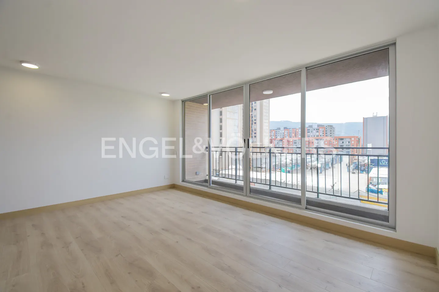 Bright, empty room with light wood floors, white walls, and large sliding glass doors to a balcony overlooking a city.