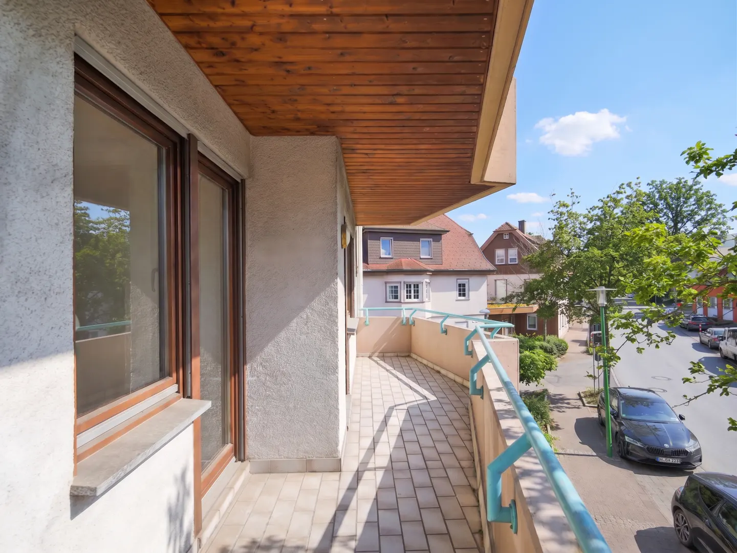 A balcony with a light beige tiled floor and a light blue railing overlooks a street with cars and trees.