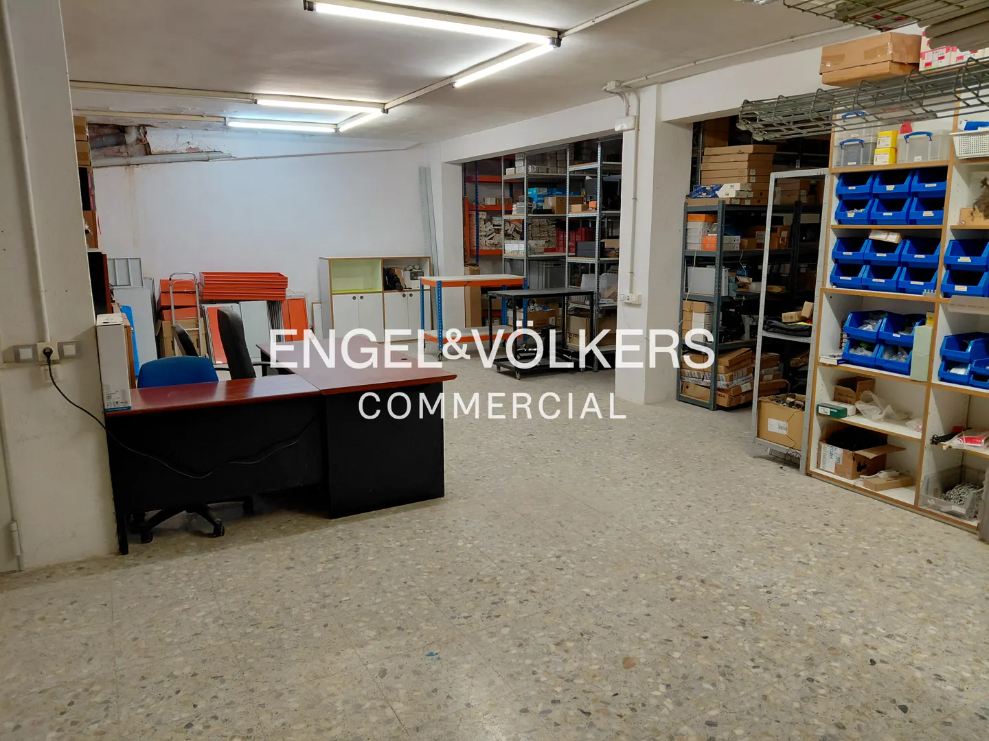 A commercial space with a desk, shelves, and storage bins. Fluorescent lights illuminate the room.