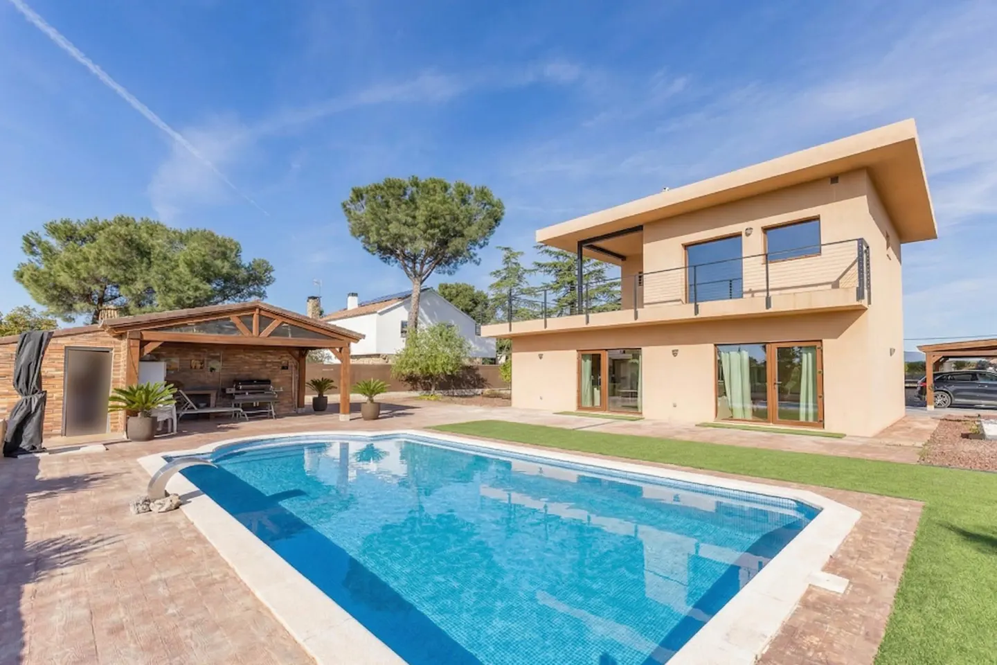 A two-story tan house with a blue swimming pool, outdoor kitchen, and a balcony on a sunny day.