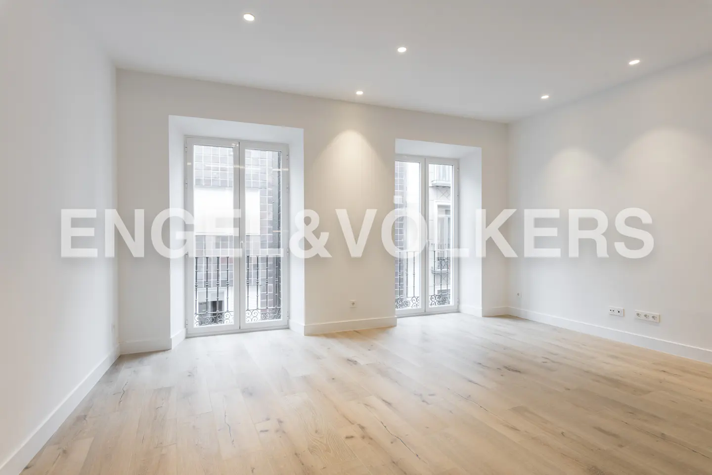 Bright, empty room with light wood floors, white walls, and two windows with black iron bars. Engel & Volkers logo overlay.