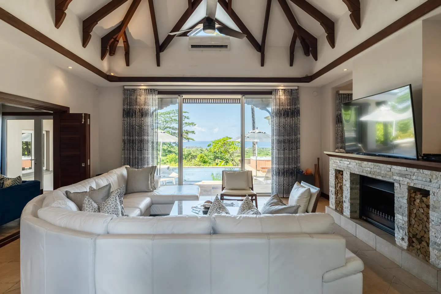 Living room with a white curved sofa, stone fireplace, and a view of a pool and ocean through sliding glass doors.