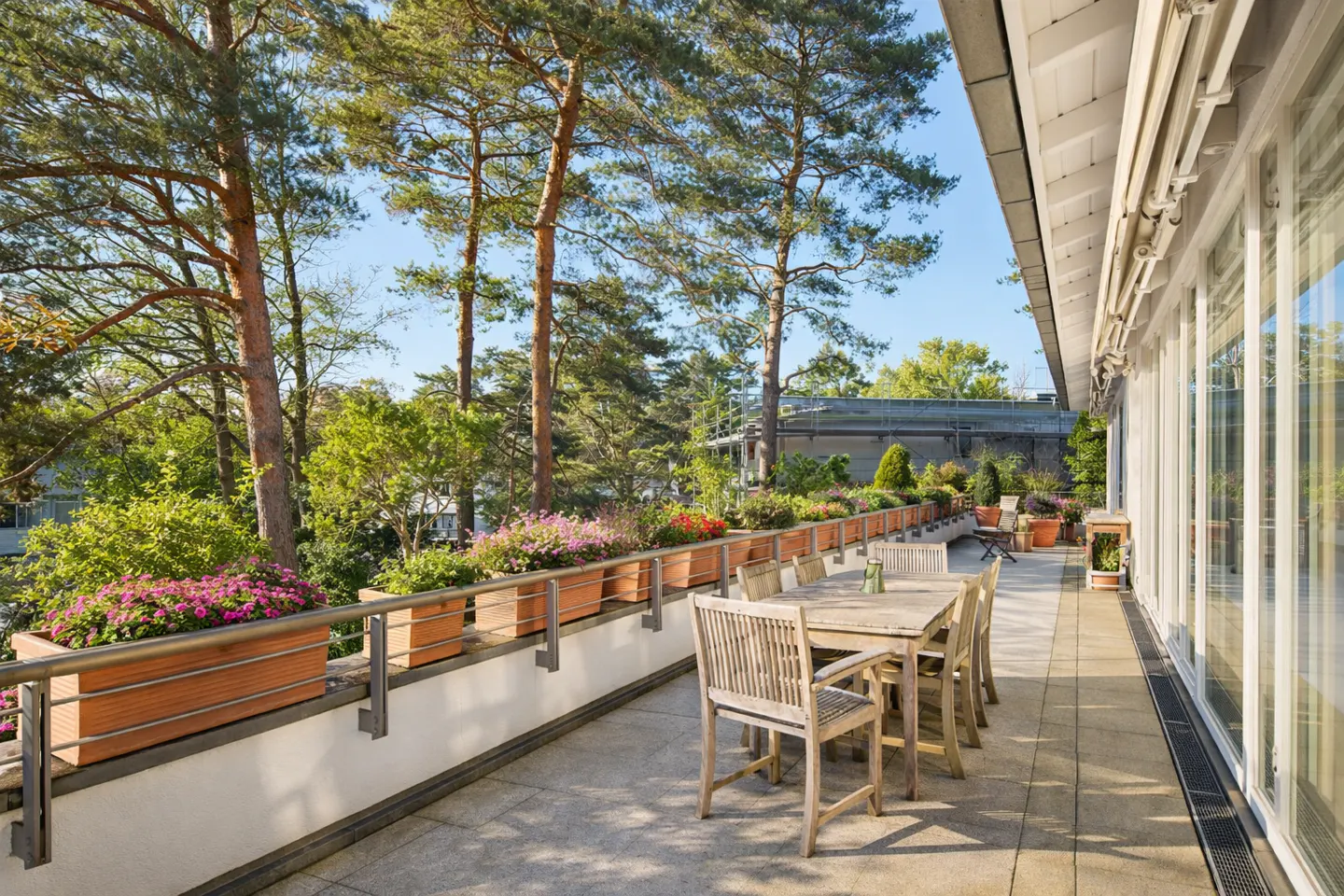 Outdoor patio with a wooden table and chairs, flower boxes with colorful flowers, and trees in the background.