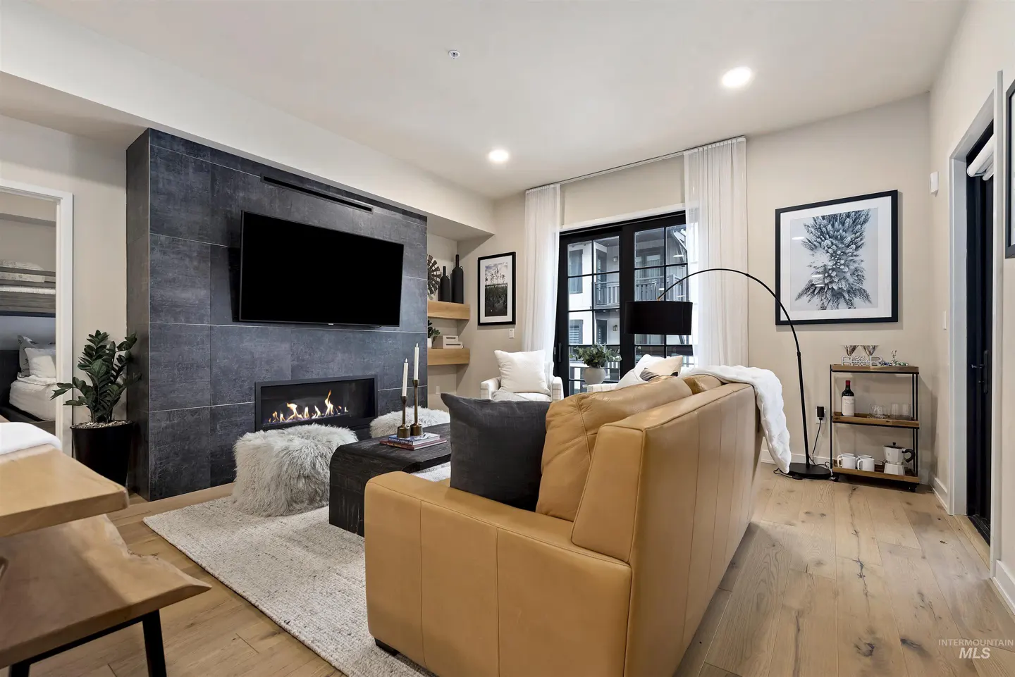 Living room with a tan leather sofa, fireplace with TV above, and a black framed window with white curtains.