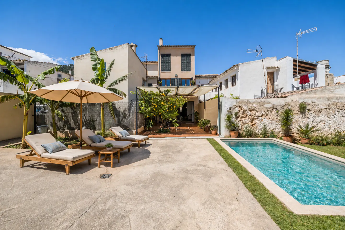 Backyard with pool, lounge chairs, and umbrella. White buildings surround the yard under a blue sky.