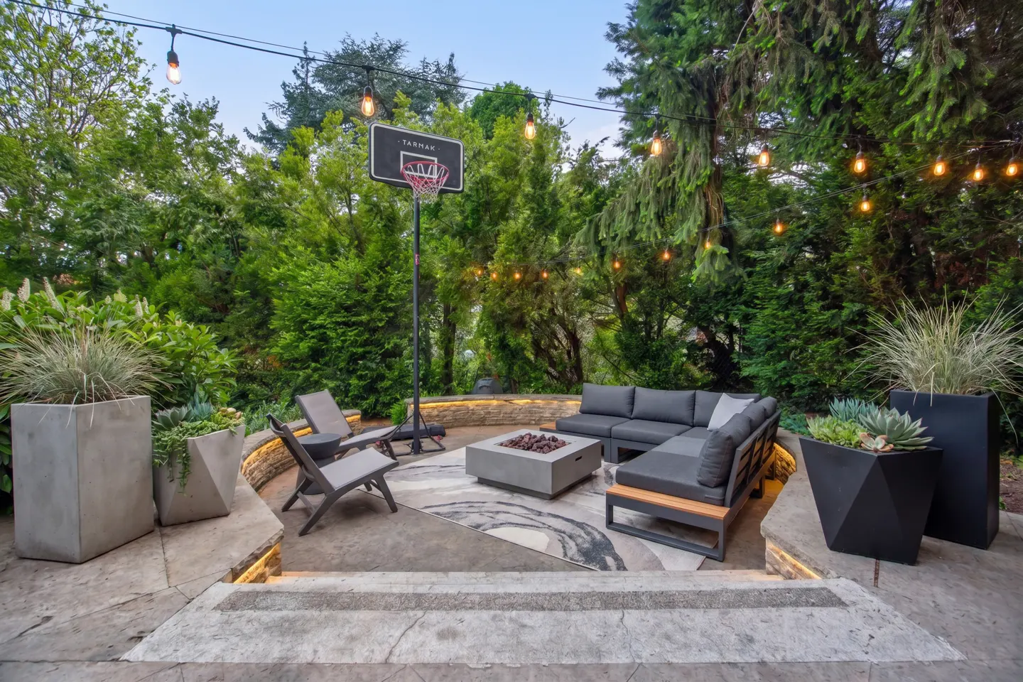 Outdoor patio with gray furniture, fire pit, and basketball hoop. String lights hang above lush green trees. Concrete planters add modern touch.