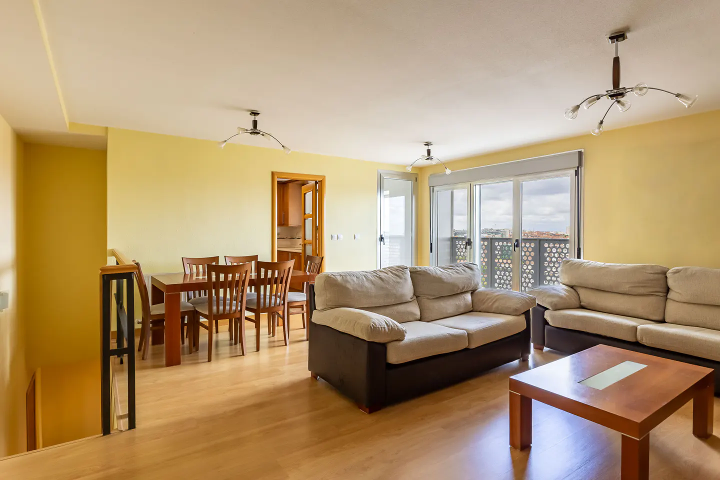 Bright living room with yellow walls, wood floors, dining table, and beige sofas. Large windows offer a city view.