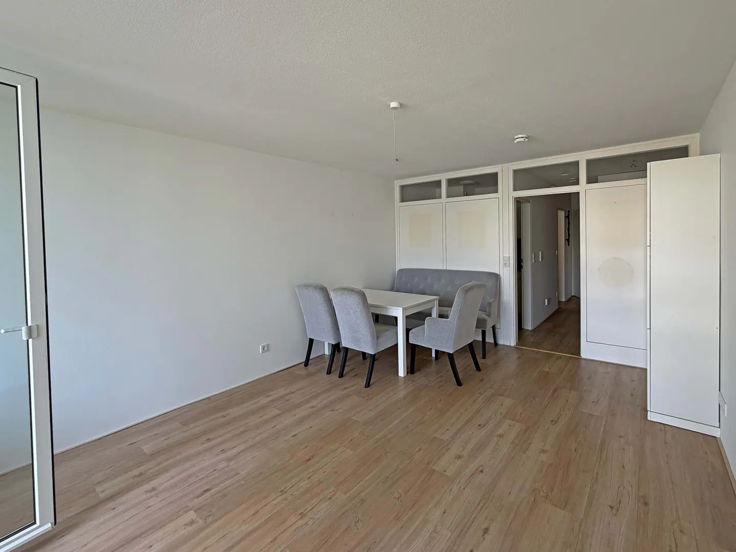 Bright, empty room with wood floors, white walls, and a dining set with gray chairs and a bench. A doorway leads to another room.