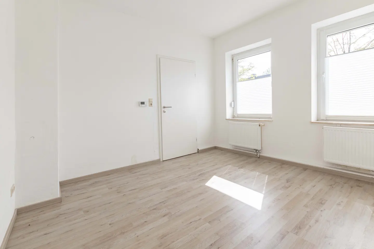 Bright, empty room with light wood floors, white walls, door, two windows with blinds, and radiators.