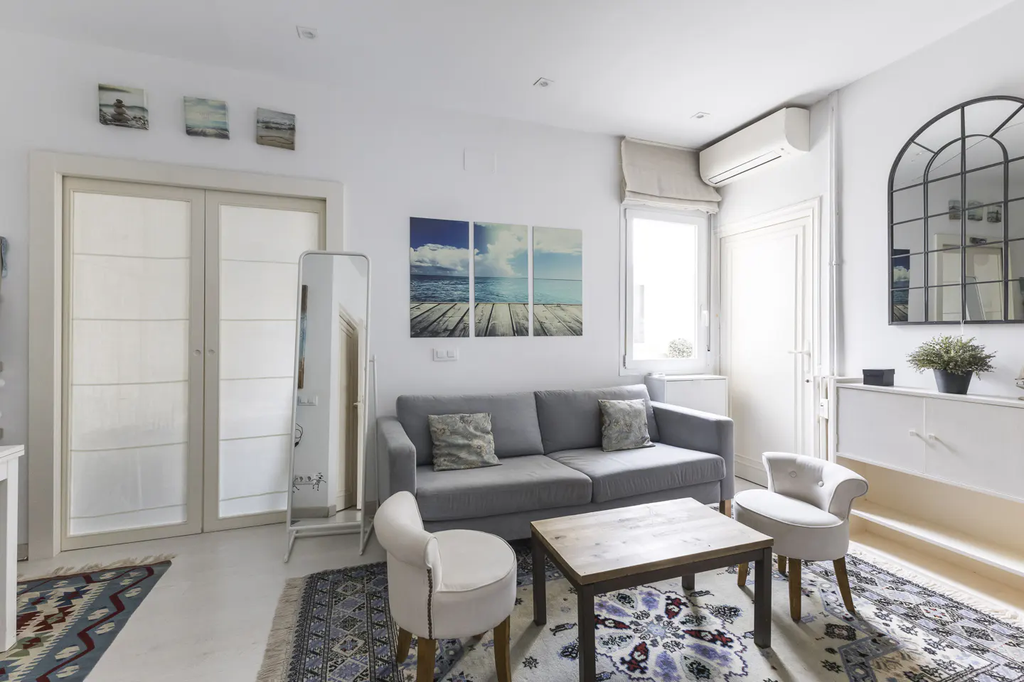 Bright living room with a gray sofa, two white chairs, and a wooden coffee table on patterned rugs. Beach-themed art adorns the white walls.