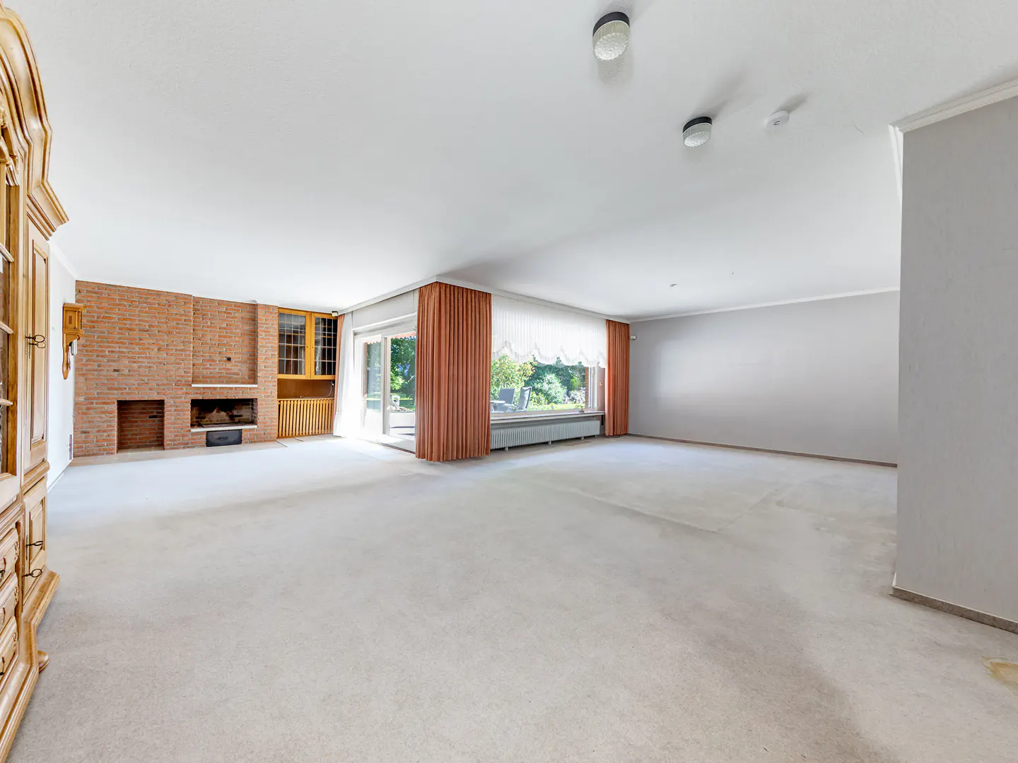 Spacious living room with white carpet, brick fireplace, wooden cabinet, and large windows with orange curtains.