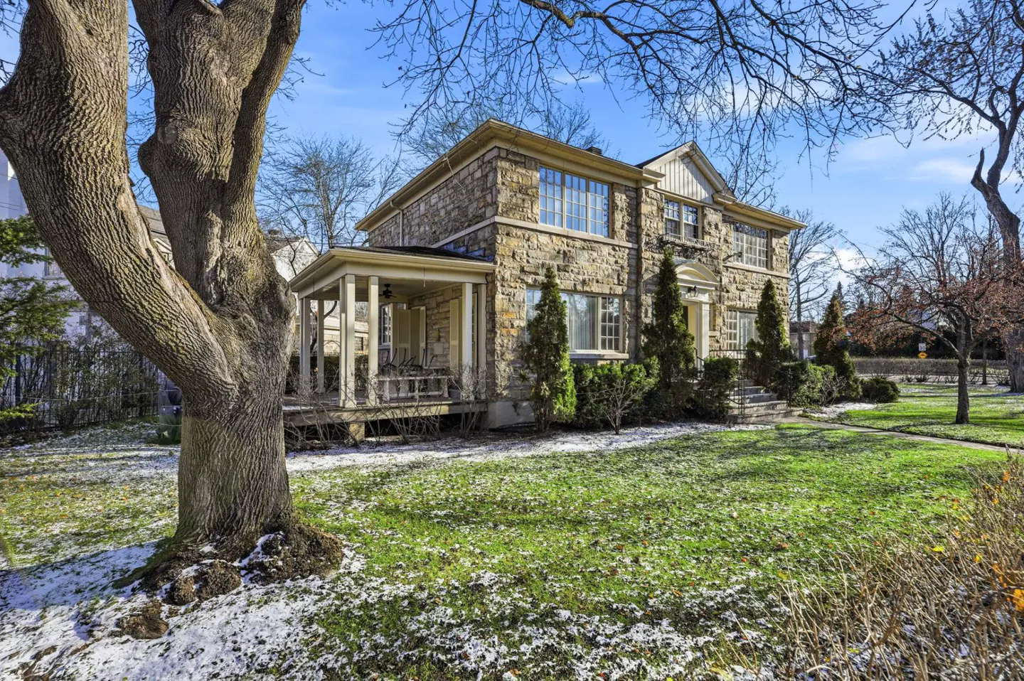Stone house with a porch and a large tree in the front yard on a sunny day with a blue sky.