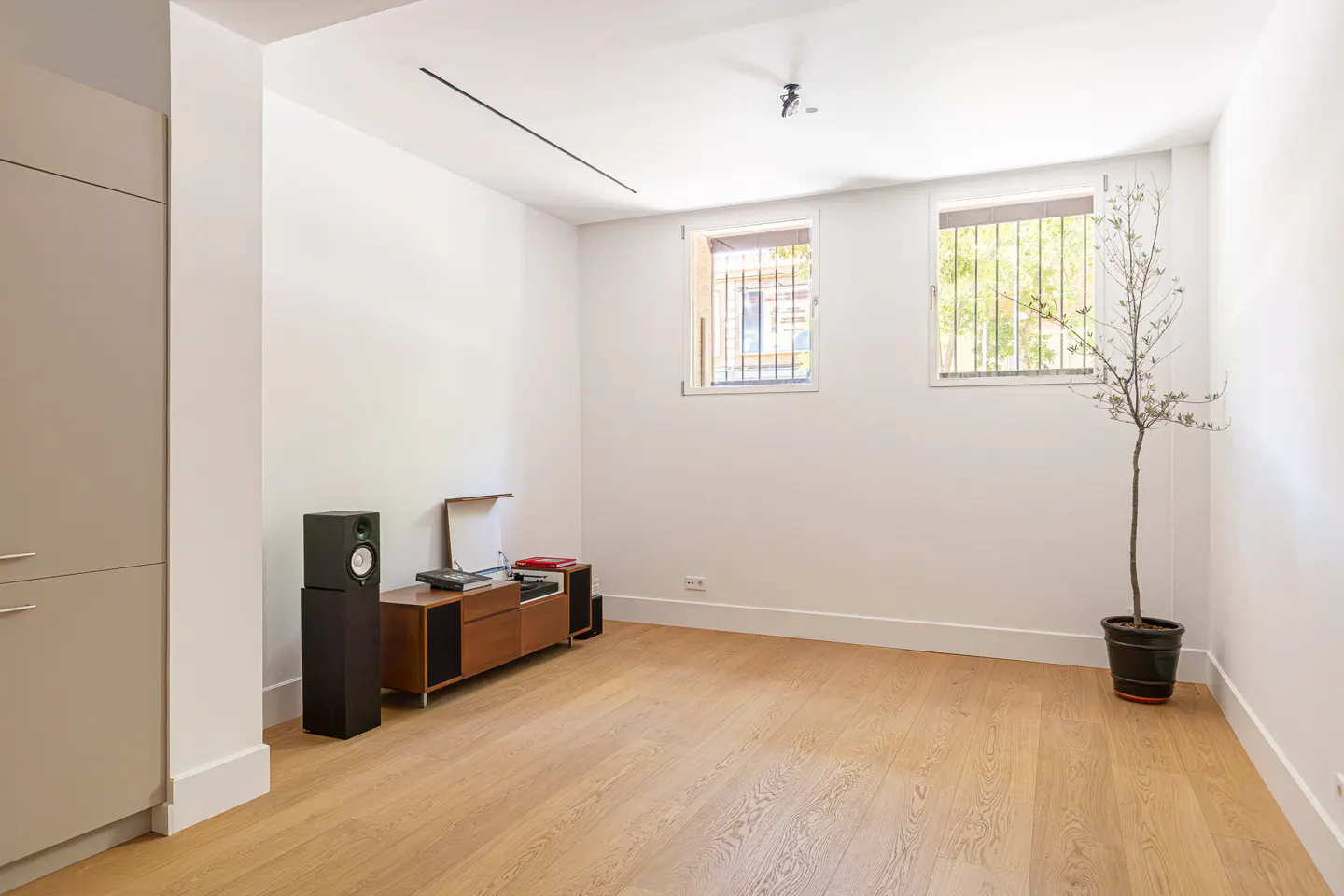 Minimalist room with light wood floors, white walls, a mid-century media console, speakers, and a potted tree by windows.