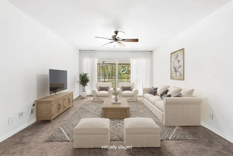 Bright living room with white walls, beige furniture, and a patterned rug. A ceiling fan hangs above a coffee table. Sliding glass doors lead to a patio.
