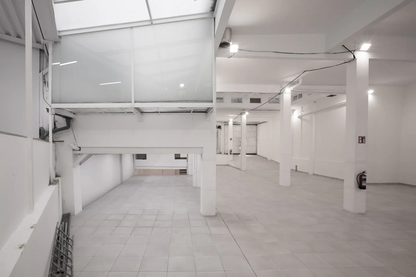 Bright, empty warehouse space with white walls, tiled floor, and support columns. Skylight visible. Fire extinguisher on a column.