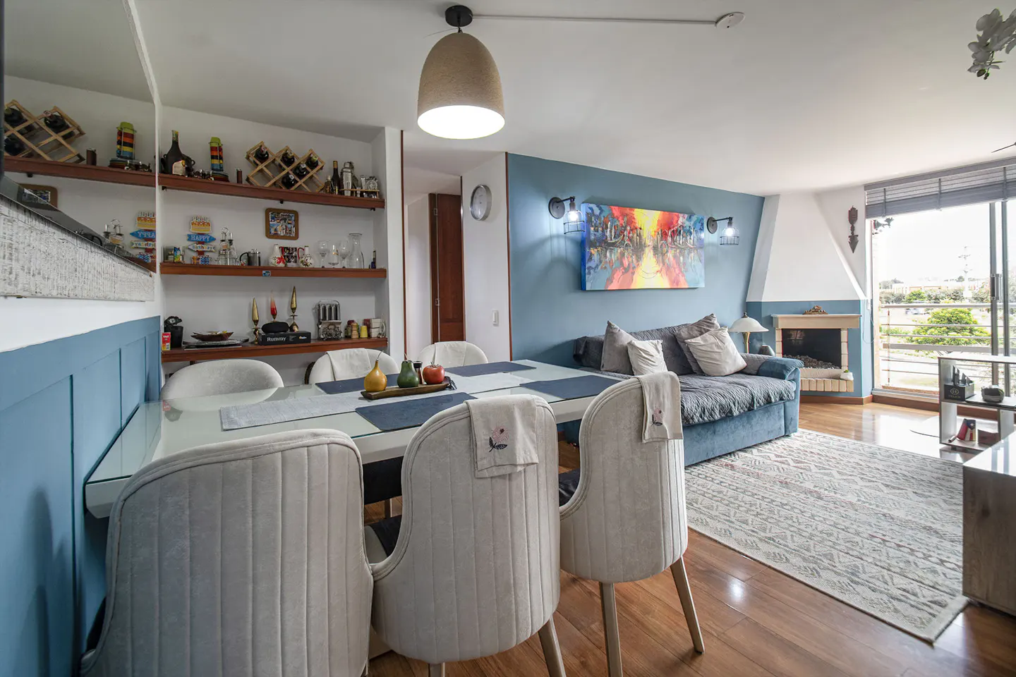 A living and dining area with a blue sofa, fireplace, and a dining table with six chairs. Shelves with decor are on the wall.
