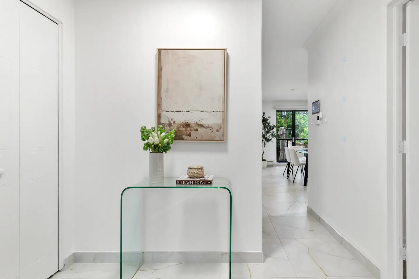 A glass console table with white tulips and a book sits against a white wall with abstract art, leading to a dining area.