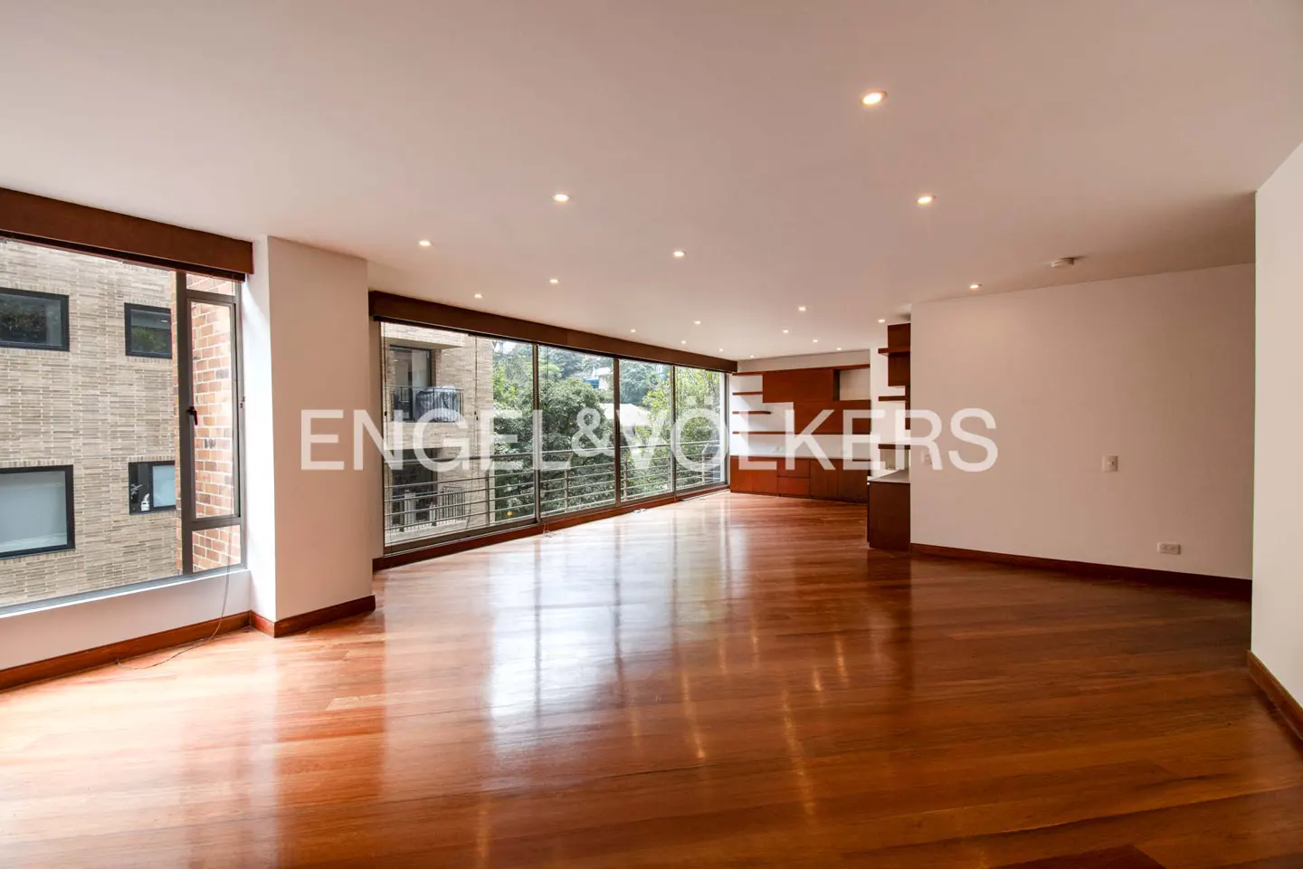 Bright, empty living room with hardwood floors, large windows, and built-in shelving. Outside view of trees and a brick building.