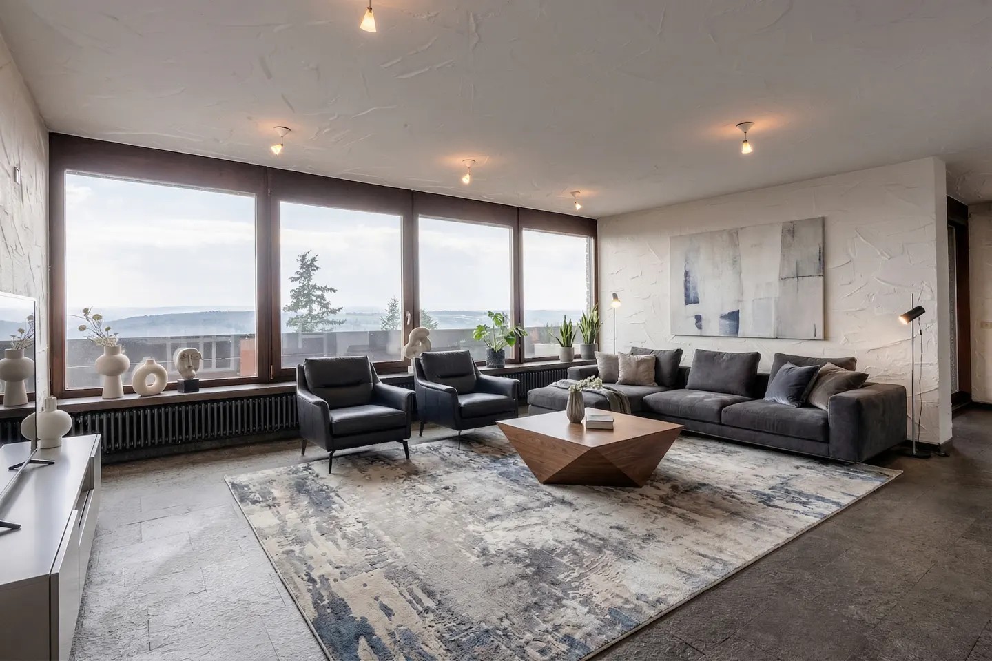 A modern living room with a gray sectional sofa, two black armchairs, and a geometric wooden coffee table on a patterned rug. Large windows offer a scenic view.