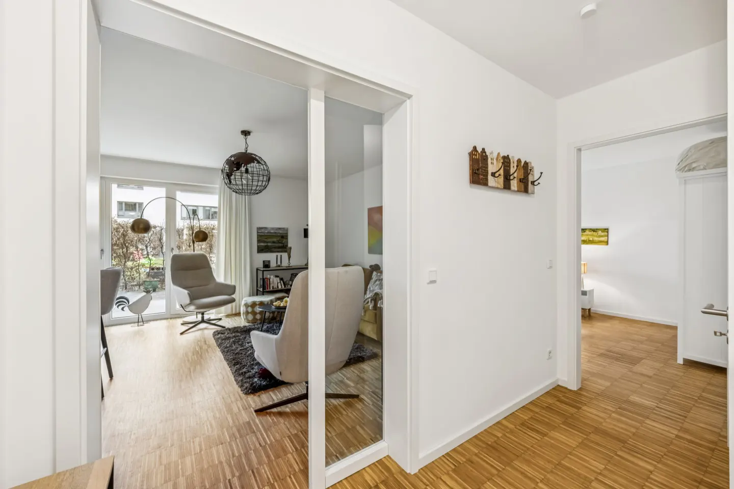 A bright hallway with wood floors leads to a living room with modern furniture and a globe pendant light.