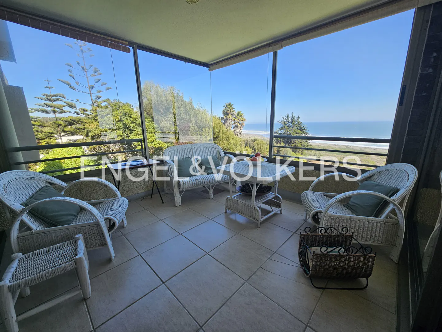 Balcony with wicker furniture overlooking the ocean on a sunny day. Engel & Volkers logo visible.