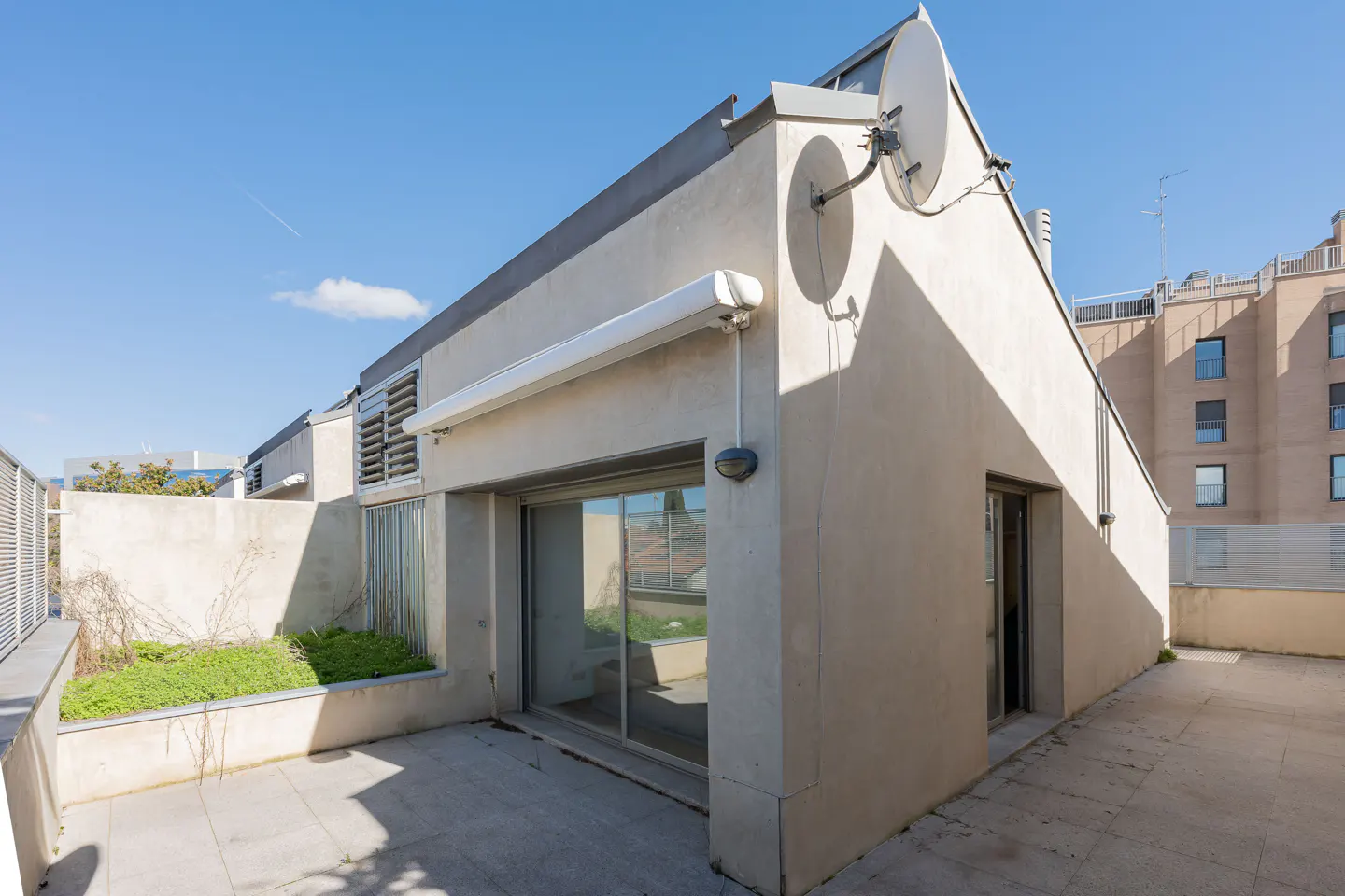 Exterior view of a modern, beige building with a flat roof, satellite dish, and a tiled patio with a small garden.