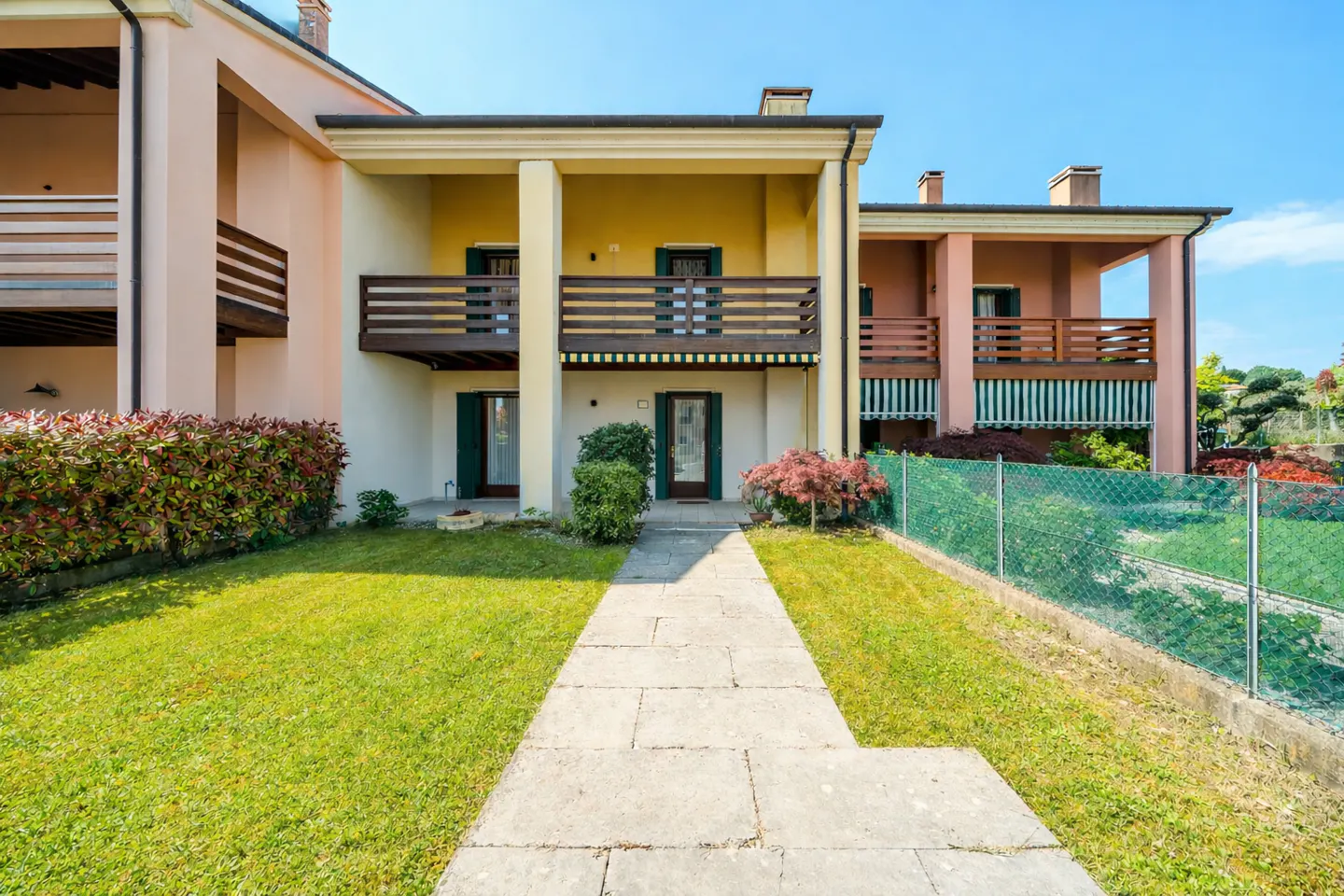 Exterior view of a two-story townhouse with a stone walkway, green lawn, and balconies. The building is painted in yellow and peach colors.