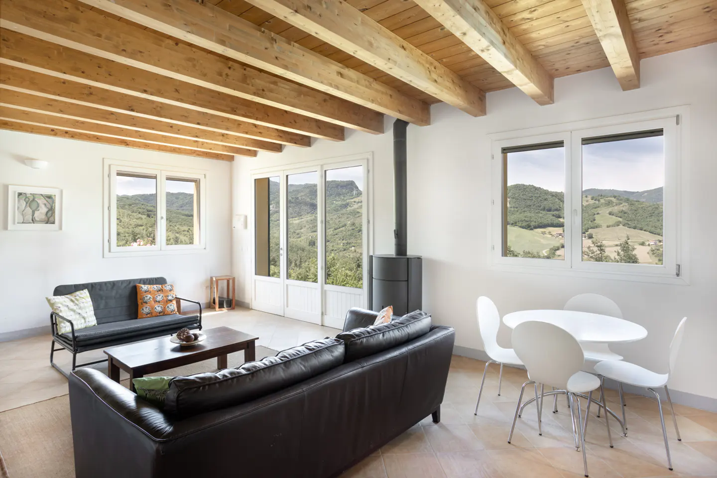 Living room with wood-beamed ceiling, black leather sofas, dining area, and large windows overlooking a mountain landscape.