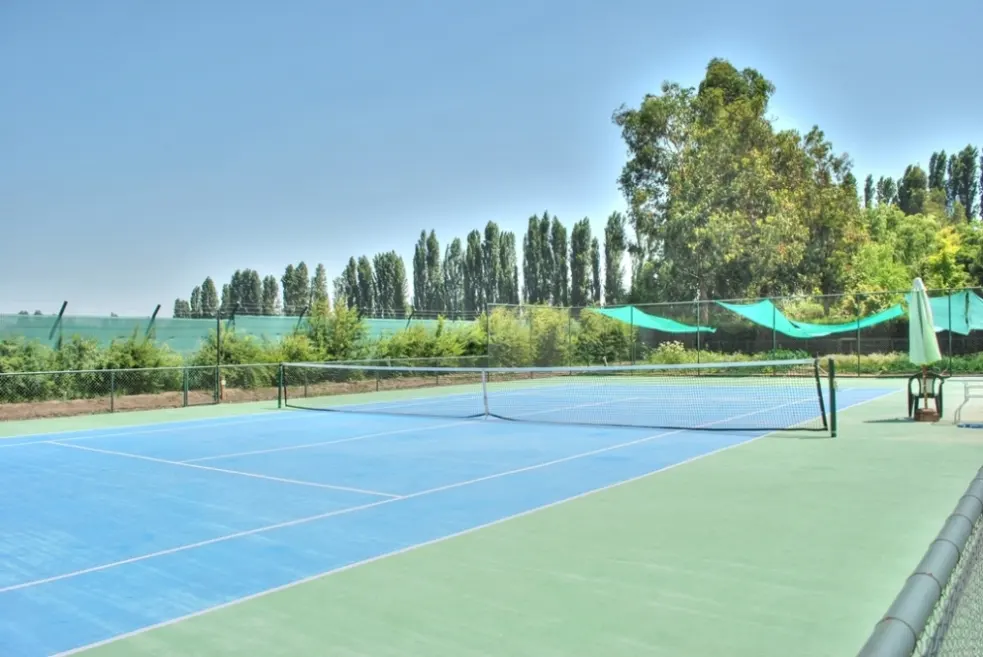 Outdoor tennis court with blue and green surface, net, and trees in the background. A chair and umbrella are on the side.