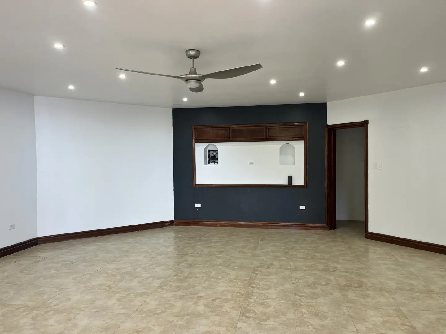 Empty room with beige tile floor, white walls, and a dark blue accent wall with wooden trim and built-in shelves. A ceiling fan is visible.