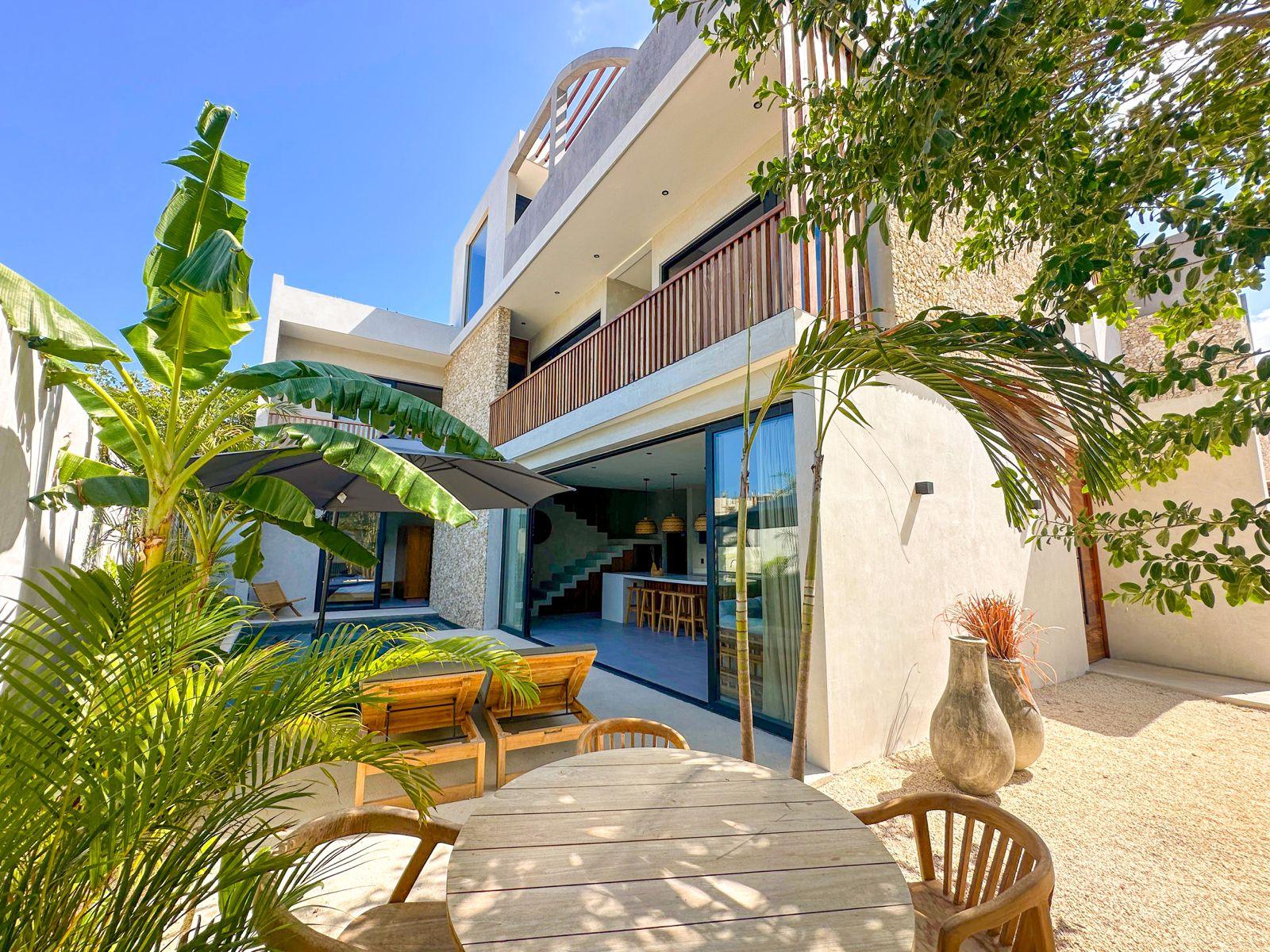 Modern villa exterior with a pool, wooden lounge chairs, patio dining set, and lush tropical plants under a blue sky.
