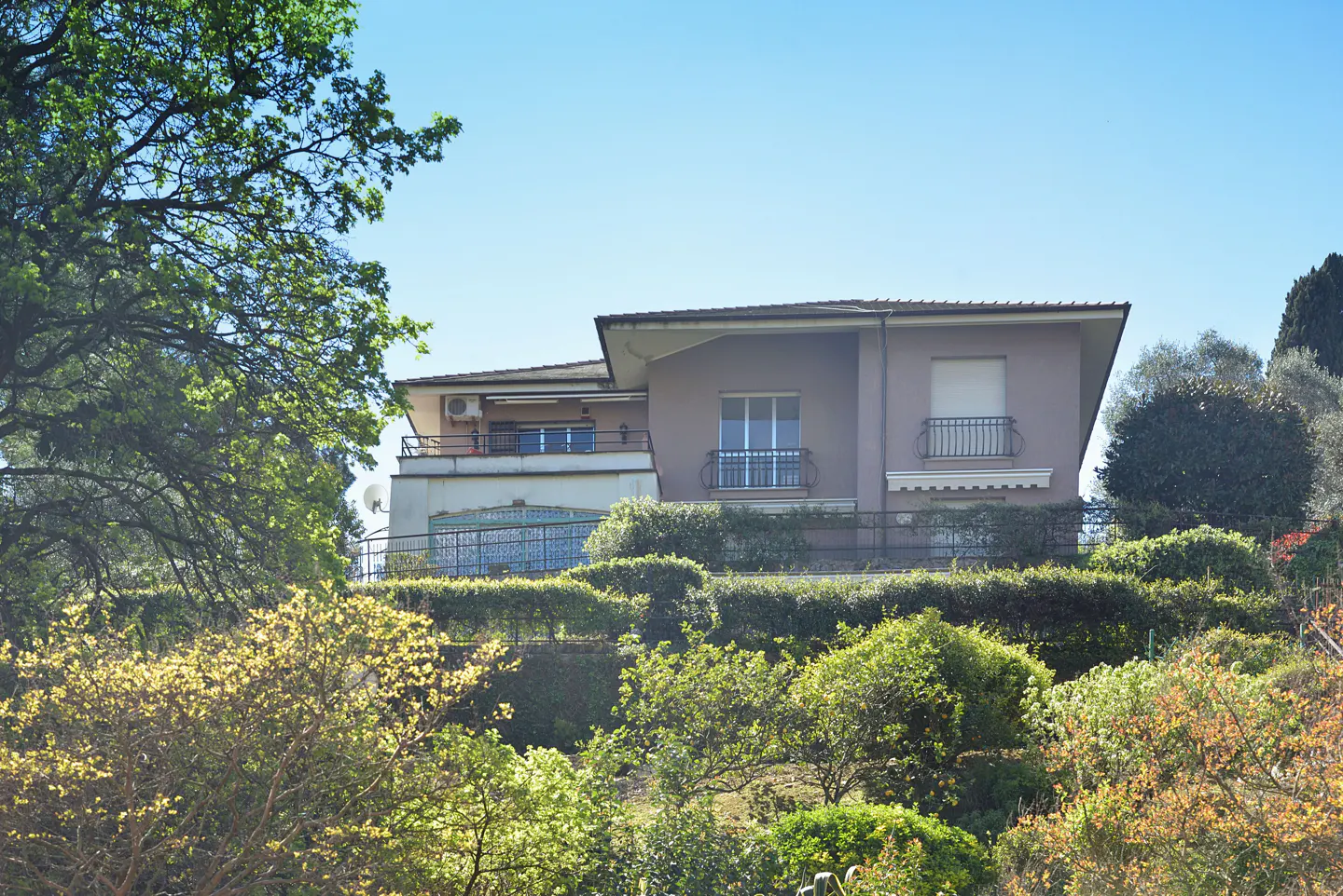 A two-story beige house with balconies sits atop a lush green hill under a clear blue sky.