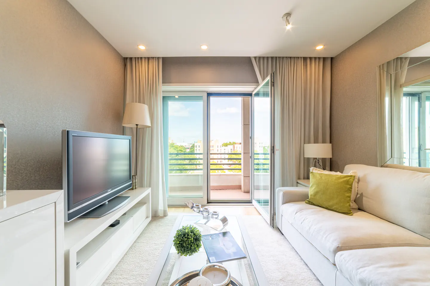 Living room with a TV, white sofa with a green pillow, and an open sliding door to a balcony.