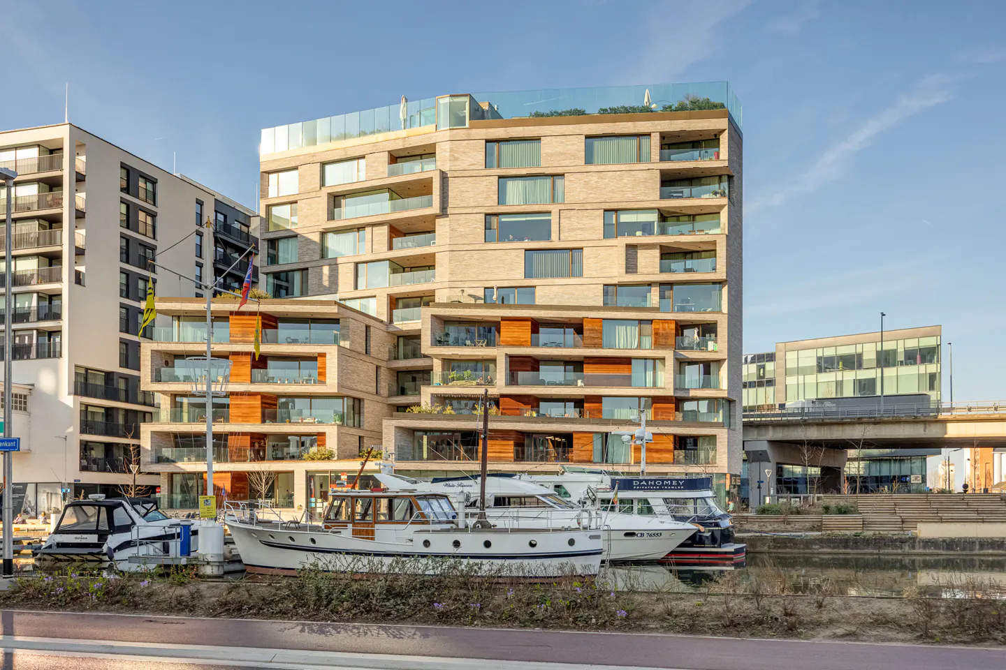 Modern apartment building with balconies overlooks a canal with docked yachts on a sunny day.