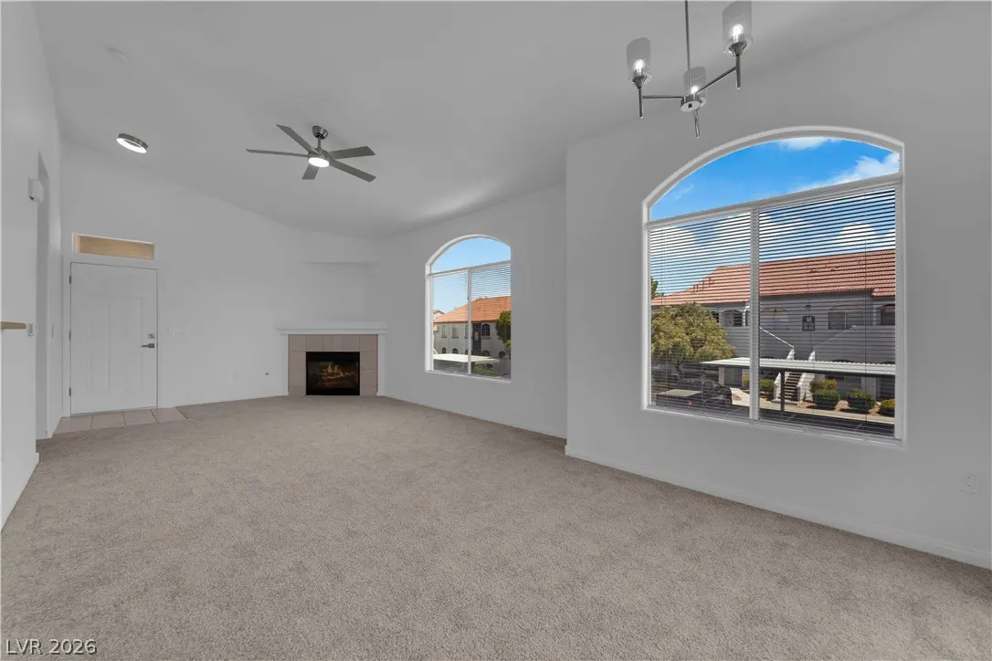 Bright, empty living room with beige carpet, white walls, fireplace, ceiling fan, and arched windows with blinds.