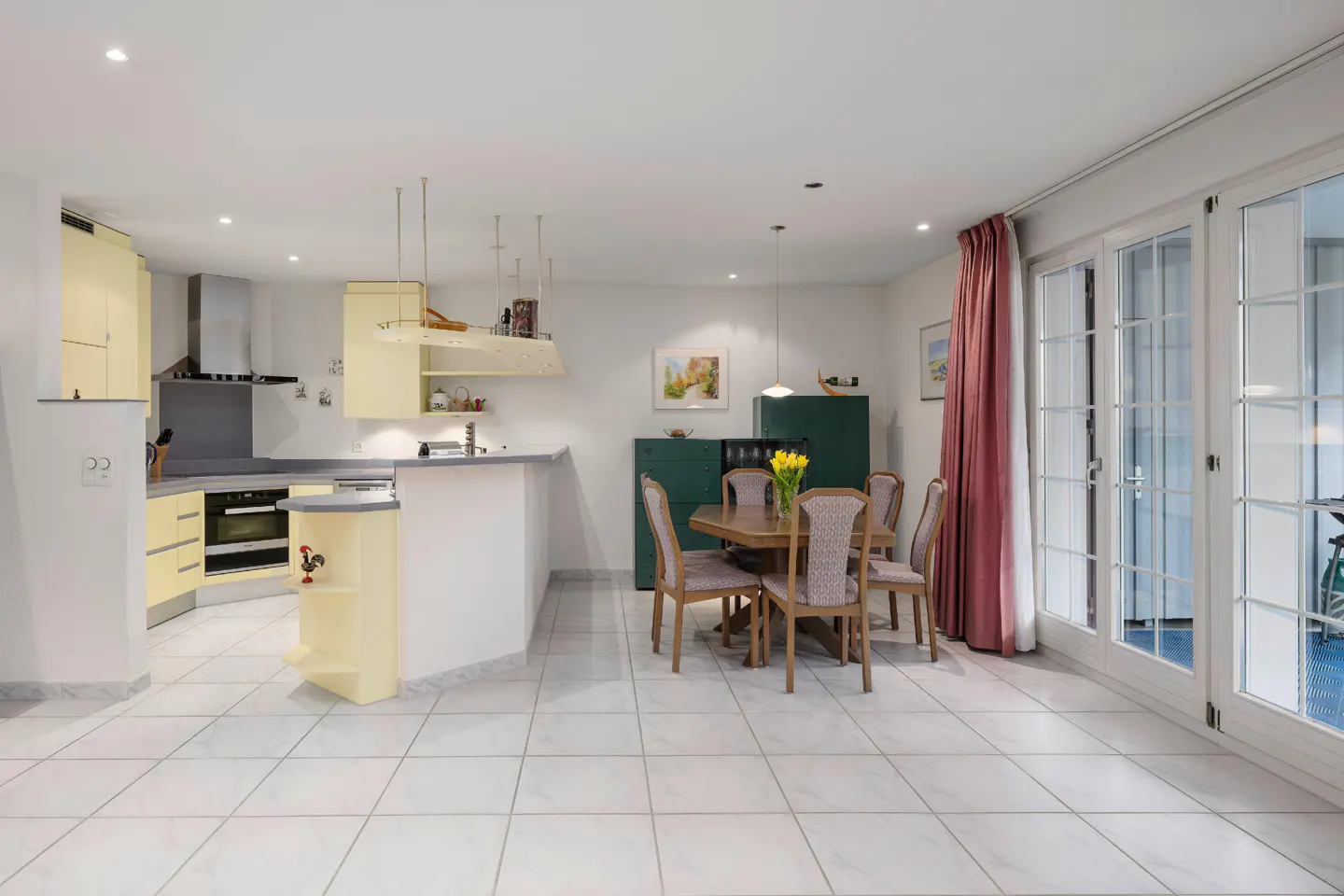 Open floor plan with yellow kitchen, dining table with tulips, and white-framed glass doors with pink curtains.