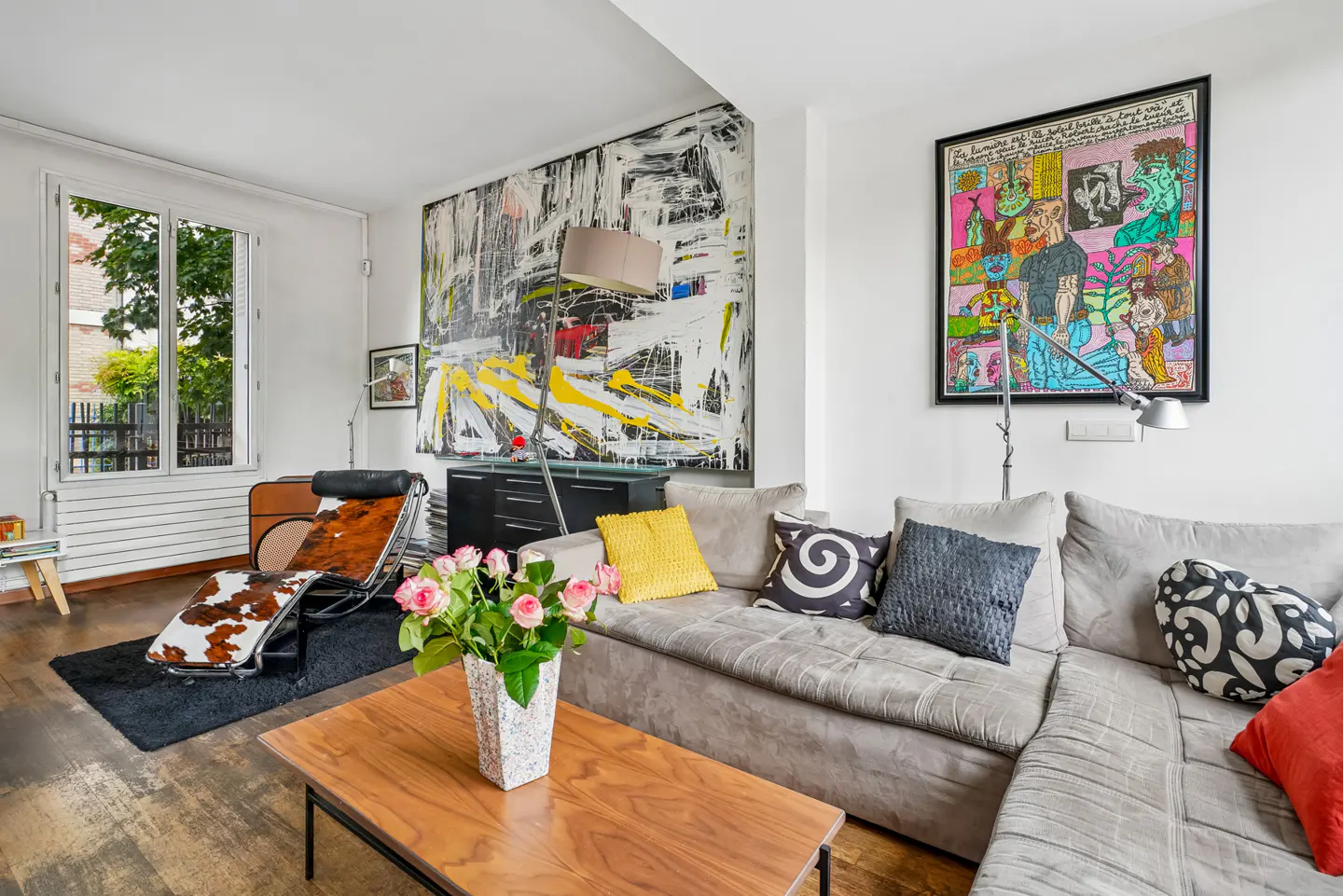 Modern living room with a grey sectional sofa, cowhide lounge chair, large abstract art, and a wooden coffee table.