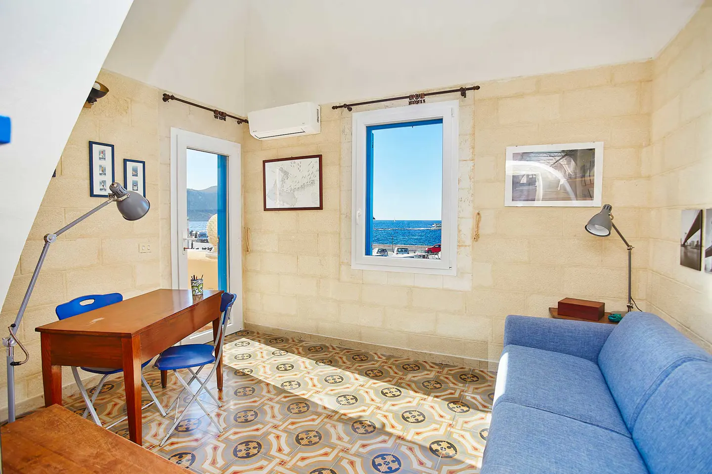 A bright room with a blue sofa, wooden desk, and chairs. A window and door offer a view of the sea. The floor has patterned tiles.