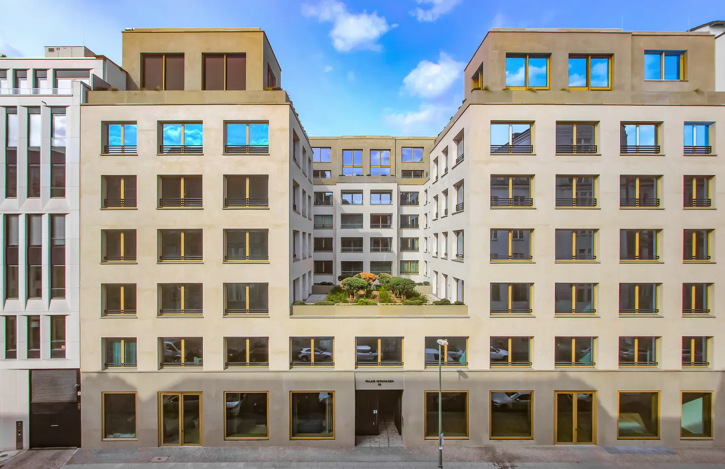 Exterior view of a modern, multi-story apartment building with many windows reflecting the blue sky. A courtyard with greenery is visible.
