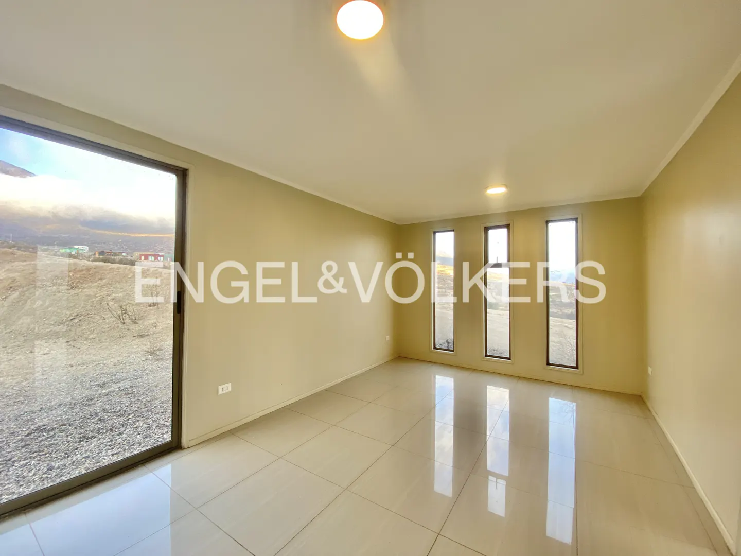 An empty room with beige walls, tile floors, and a large sliding glass door. Three vertical windows let in natural light. The Engel & Völkers logo is superimposed.