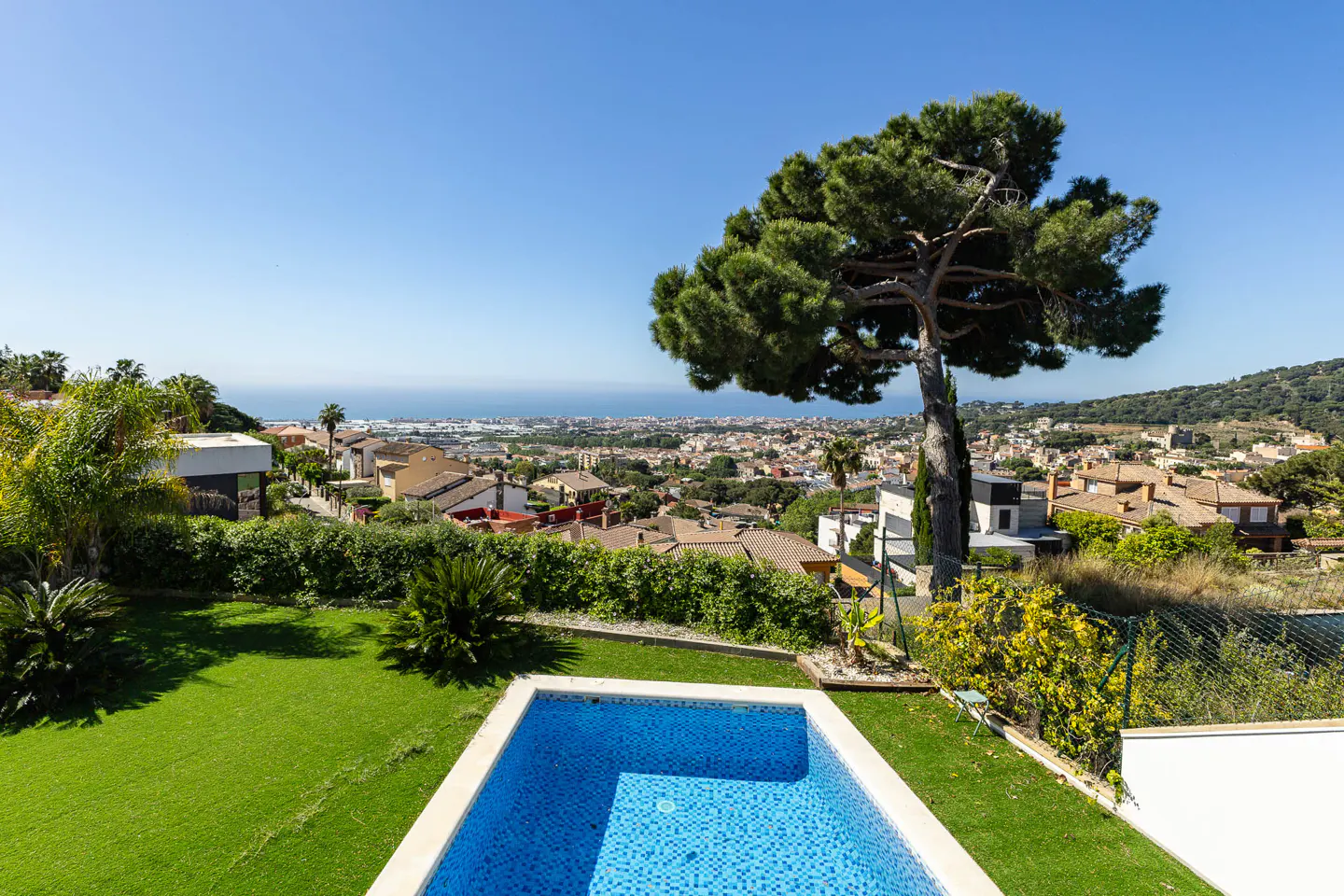 A backyard with a blue tiled pool, green grass, and a large tree overlooking a town and the ocean under a clear blue sky.